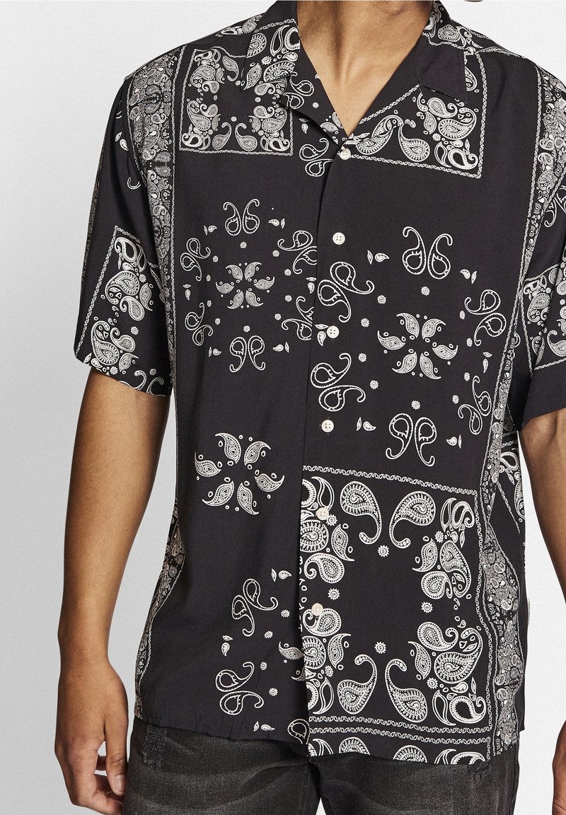 Chemise noire à manches courtes boutonnée avec motifs bandana blancs de style paisley et floraux portée par une personne du cou aux hanches.