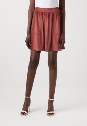 Kvinde iført en rust-rød, knælange nederdel med lappelommer og hvide, åbne høje hæle-sandaler på hvid baggrund.