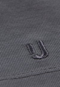 Szary, dzianinowy materiał o fakturowanej powierzchni; na pierwszym planie widnieje ciemnoniebieska haftowana litera "U".