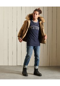 Chaqueta acolchada color marrón con capucha de pelo sintético, camiseta azul marino con el texto "PISTONS", jeans de color azul claro y botas negras con cordones. Entorno interior.