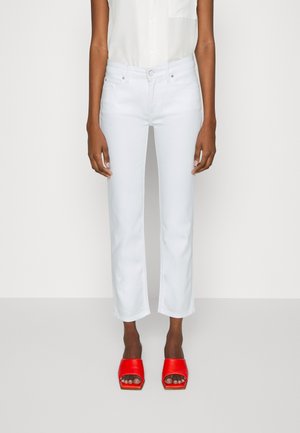 Femme portant un jean blanc court, un haut blanc sans manches et des sandales rouges à talons ouverts, debout devant un fond uni.