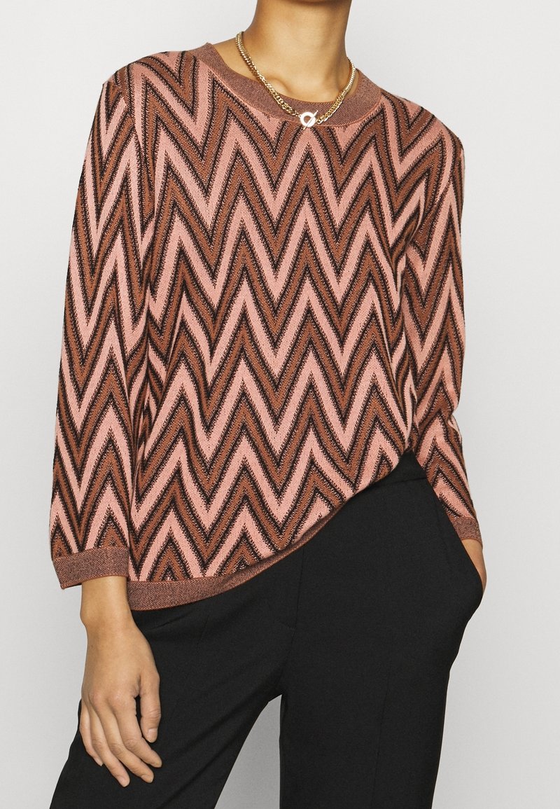 Różowo-czarny sweter z wzorem w zygzaku, dzianinowy, z długimi rękawami i okrągłym dekoltem, zestawiony z czarnymi spodniami. Widoczny złoty naszyjnik z łańcuchem.