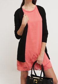 Černý cardigan přes korálové šaty s krajkovým lemem. Šaty mají hladký povrch a kulatý výstřih, doplněné černou kabelkou.