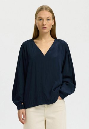 Kvinne med rett blond hår iført en løs marineblå langermet bluse og lysebeige bukser, står foran en ensfarget hvit bakgrunn.