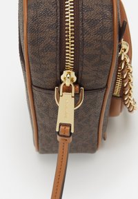 Borsa in pelle marrone con motivo, zip dorata e fibbia della tracolla incisa con "MICHAEL KORS" su sfondo bianco.