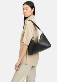 Sac à main en cuir noir avec une texture lisse, un design courbé et une bandoulière ajustable, tenu par une personne portant une chemise rayée beige clair.