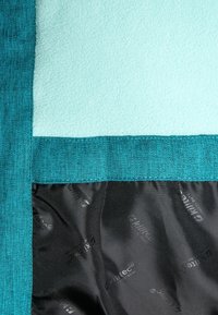 Fleecefilt med en mjuk ljusblå yta, kantad av strukturerad teal-tyg; har ett svart innerfoder med ett upprepande logotypmönster.