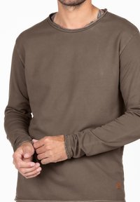 Camicia marrone a maniche lunghe, realizzata in morbido tessuto, presenta un colletto rotondo, polsini sfrangiati e una piccola etichetta con logo in basso a destra.