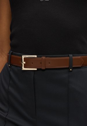 Brun læderbælte med en guldfarvet metalspænde, der har et rektangulært design og et logo-tag. Bæltet bæres over mørke stofbukser.