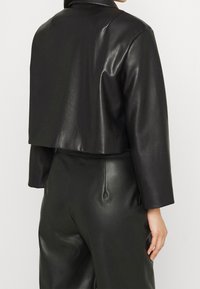 Giacca nera in ecopelle con finitura lucida, colletto a revers e maniche lunghe. È abbinata a pantaloni a vita alta coordinati.