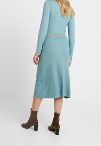 Stickad klänning i medellång längd i ljus teal med långa ärmar och diagonala texturerade mönster. Har en åtsydd midja och subtila spetsdetaljer.