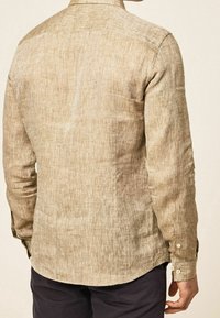 Chemise en lin beige clair avec une surface texturée, des poignets à boutons et un ourlet arrondi, présentant un design simple et minimaliste.