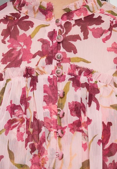 Blouse fleurie rose clair, ornée de grandes fleurs bordeaux, avec un col à volants et des boutons roses à détails argentés sur le devant.