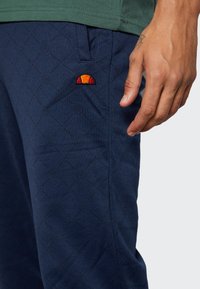 Pantalones azul marino con un patrón de diamantes texturizado, que presenta un logo bordado en naranja y rojo en el bolsillo. Tela suave y diseño ajustado.
