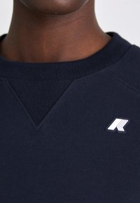 Felpa blu navy con colletto a girocollo ribattuto e dettagli di cucitura triangolari sul colletto. Presenta un piccolo logo bianco sul petto.