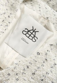 Balts strukturēts audums ar melnām šķipsnām, ar gludu iekšējo oderi. Ir pievienota etiķete ar uzrakstu "KAREN BY SIMONSEN".