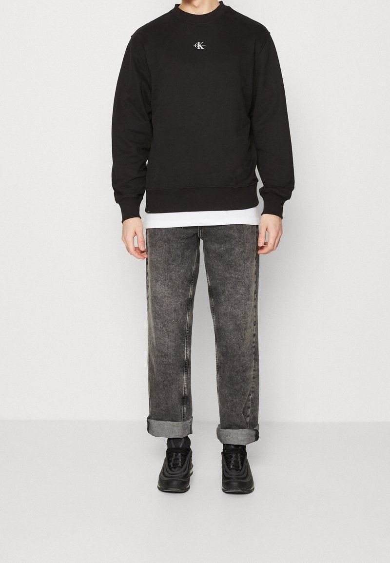 Personne portant un sweat-shirt Calvin Klein noir, un sous-pull blanc, un jean noir délavé retroussé et des baskets noires, debout devant un fond uni.
