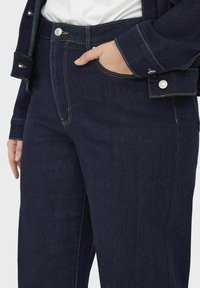 Mörkblå denimjeans med gula sömmar och en framficka; har en knappstängning och är stylad med en matchande jacka.