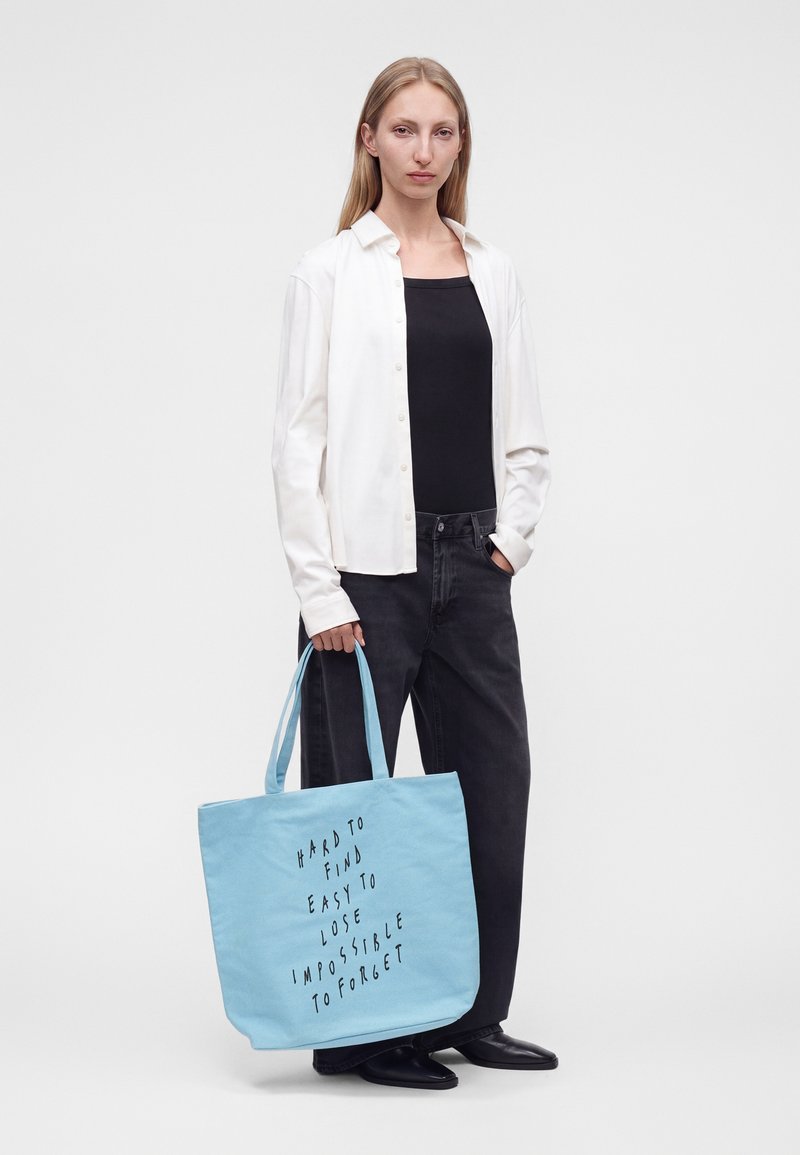 Femme aux longs cheveux blonds portant une chemise blanche, un top noir et un pantalon noir, tenant un sac fourre-tout bleu clair avec un texte noir.