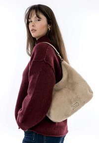 Sac à main en fausse fourrure beige avec une texture douce et une forme arrondie, présentant une sangle contrastante et des détails en métal.