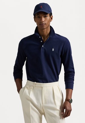 Navy long-sleeve polo shirt with a three-button placket and white embroidered logo, paired with cream pleated trousers and a matching cap.