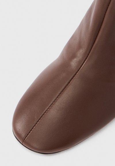 Bottine en cuir marron à texture lisse, bout arrondi et détail de couture sur le côté. La semelle est noire et plate.