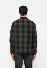 Mann iført en kviltet grønn og svart rutet jakke og mørke jeans, vendt mot en enkel hvit vegg.