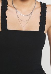 Frau, die ein schwarzes ärmelloses Oberteil mit Rüschen-Trägern und geschichteten silbernen Halsketten auf barer Haut trägt.