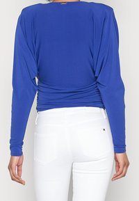 Blå långärmad topp med samlad ryggdetalj, matchad med vita tighta byxor; slät tyg med lätt textur.