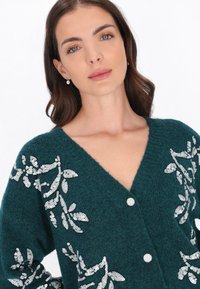 Cardigan de punto en color verde azulado con escote en V, decorado con patrones florales de lentejuelas blancas y botones redondos en la parte delantera. Textura suave y mangas abiertas.