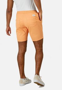 Pantalones cortos naranja con una cinturilla elástica y dos bolsillos traseros. Llevados con zapatillas blancas y beige. Detalles de textura y color visibles.