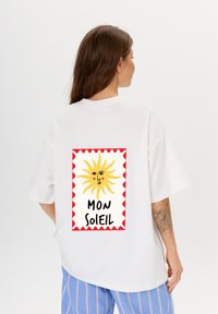Weißes übergroßes T-Shirt mit einem Grafikdesign auf der Rückseite, das eine gelbe Sonne mit rotem Rand und dem Text "MON SOLEIL" in fettgedruckten schwarzen Buchstaben zeigt.