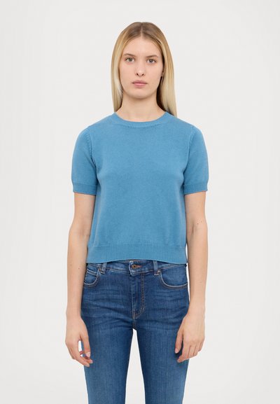 Lys blå strikket kortermet genser med ribbestrikket hals og kant, paret med mørkeblå jeans, vist mot en nøytral bakgrunn.