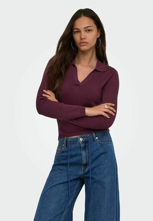 Jeune femme aux longs cheveux bruns portant un pull violet côtelé et un jean bleu, debout, les bras croisés, devant un fond uni.