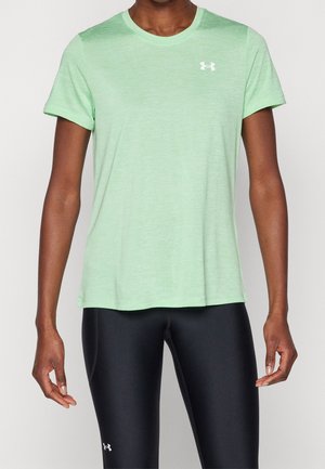 Osoba ubrana w jasnozieloną koszulkę Under Armour z krótkim rękawem oraz czarne dopasowane legginsy z logo Under Armour na lewej części uda.