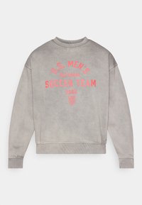 Pelēks džemperis ar ribotu aproču un apakšmalu. Priekšpusē sarkani burti: "U.S. VĪRIEŠU NACIONĀLĀ FUTBOLA KOMANDA 2026" ar logotipa akcentiem.