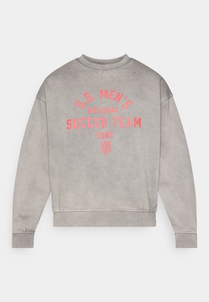 Harmaa collegepaita, jossa resorit hihansuissa ja helmassa. Edessä punainen teksti: "U.S. MEN'S NATIONAL SOCCER TEAM 2026" ja logokoristeita.