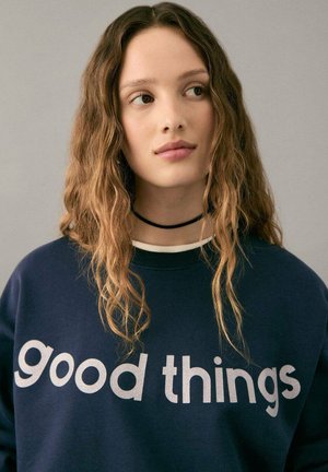Tamnoplava majica s kapuljačom s srebrnim natpisom "good things" na prednjoj strani. Model ima valovitu kosu i nosi crni choker. Jednostavna pozadina.