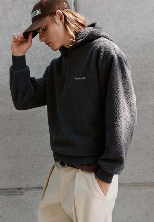 Persoon draagt een donkergrijze, gestructureerde hoodie en beige broek, stelt een bruine pet af, staand tegen een eenvoudige betonnen muur.