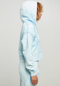 Lys blå, kort hoodie med tie-dye-mønster. Har hette og ribbestrikkede mansjetter. Stoffet ser mykt ut med en avslappet passform.