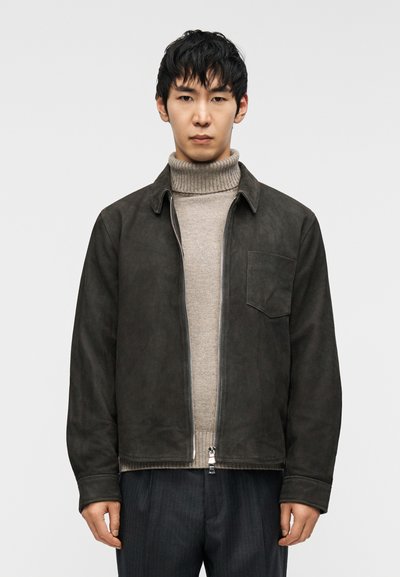 Brown suede jacket with a collar, front zipper, and pocket. Worn over a light gray turtleneck. Dark striped trousers.