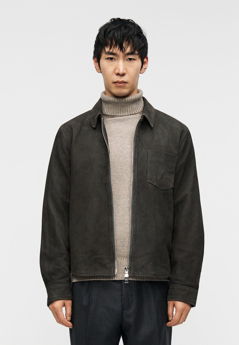 Jachetă maro din piele întoarsă, cu guler, fermoar frontal și buzunar. Poartă-o peste un helancă gri deschis. Pantaloni în dungi închise la culoare.