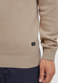 Beige strikket sweater med ribbet kant og manchetter. Lille sort label nær bunden med mærkenavn. Glat tekstur, medium tykkelse.