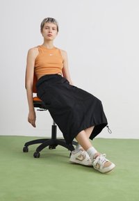 Top arancione senza maniche, gonna nera di lunghezza media con coulisse, sneakers bianche con dettagli beige, seduta su una sedia ergonomica nera.