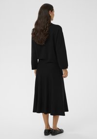 Naine, kellel on pikad lainelised juuksed, kannab musta pikakäiselist toppi, musta midi-seelikut ja musti loafereid, seistes valge tausta poole seljaga.