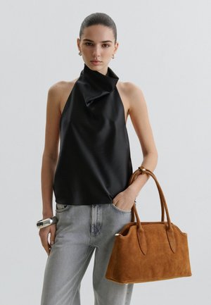 Vrouw in zwart mouwloos haltertopje en grijze spijkerbroek, met een bruine suède handtas, één hand in de zak.