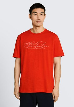 Mężczyzna noszący jasnoczerwoną koszulkę z krótkim rękawem z podwiniętym mankietem i jasnym graficznym napisem na piersi, stojący na jednolitym tle.