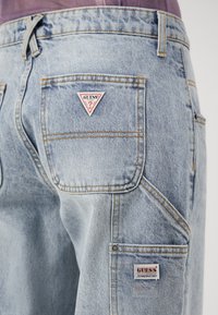 Ljusblå denimjeans med kontrasterande sömmar, som har en triangulär "GUESS"-logotyp på den högra bakfickan och en annan märkesetikett.