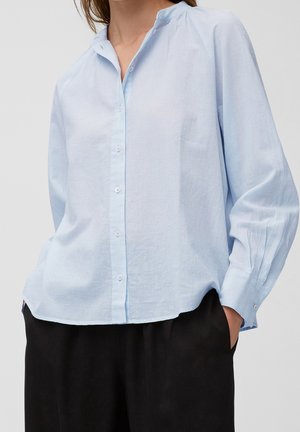 Femme portant une chemise bleu clair à manches longues avec col mandarin, légèrement rentrée dans un pantalon noir, les mains dans les poches.