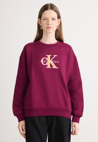 Bordový sveter s okrúhlym výstrihom a dlhými rukávmi. Obsahuje vyšívané logo "Calvin Klein" a "CK" s odtieňmi zlata.